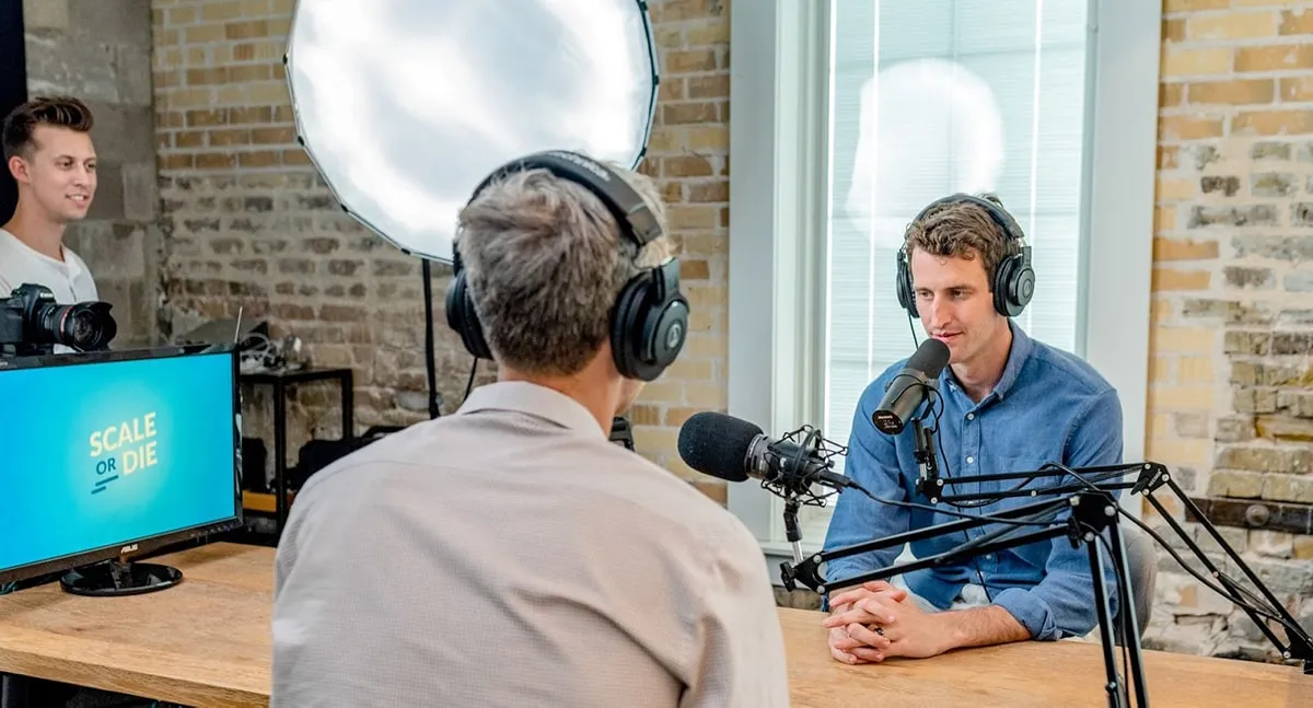 Podcast host interviewing a guest at microphones in a brick-walled studio