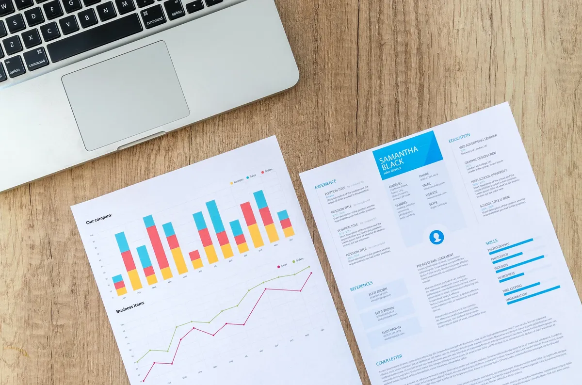 Printed charts and a resume next to a laptop on a wooden desk