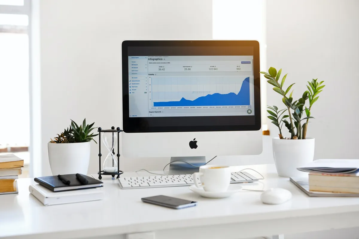 Desk with an iMac showing a marketing analytics chart, a notebook and a coffee cup