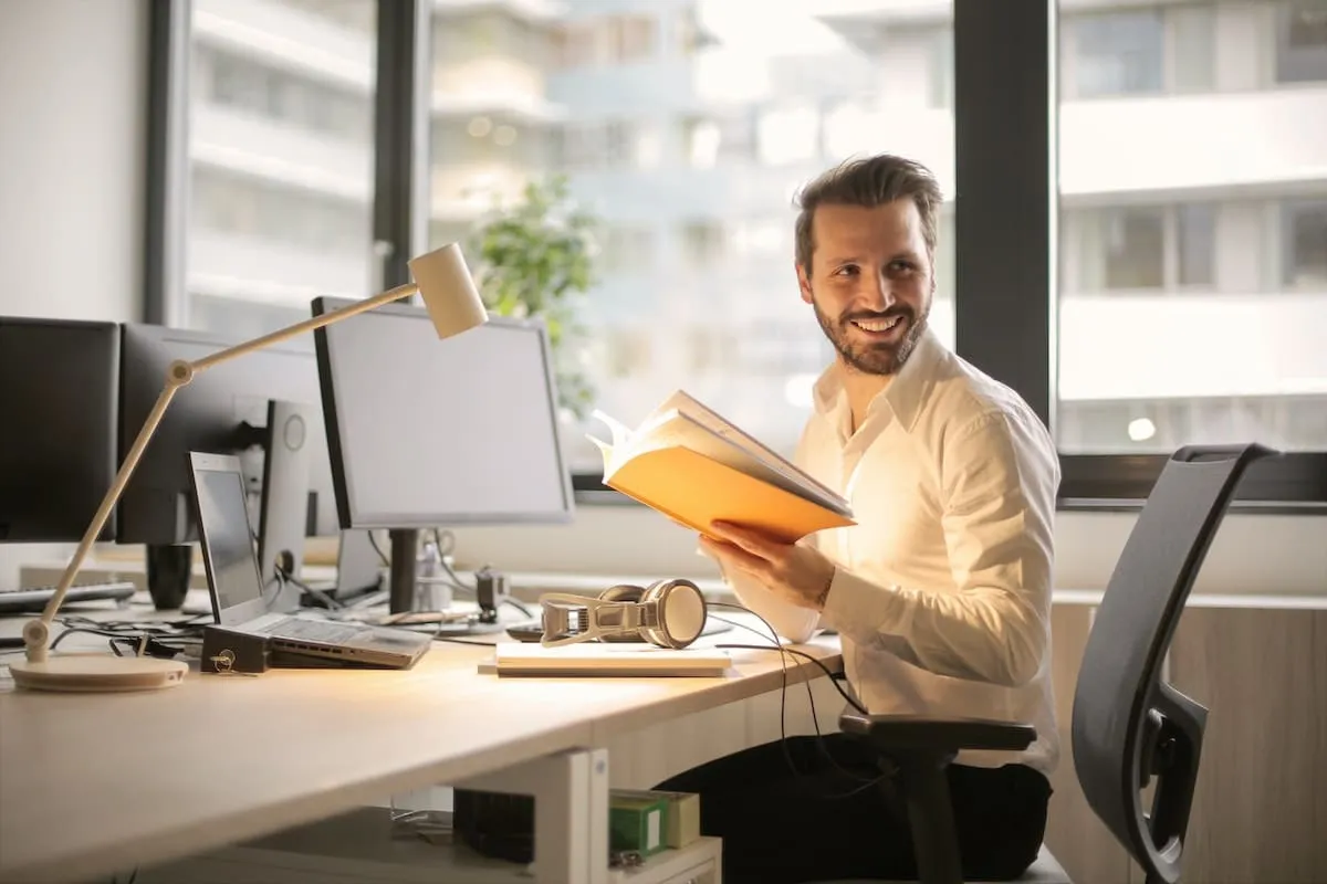 A man at a desk with two monitors and a laptop, reading through a notebook