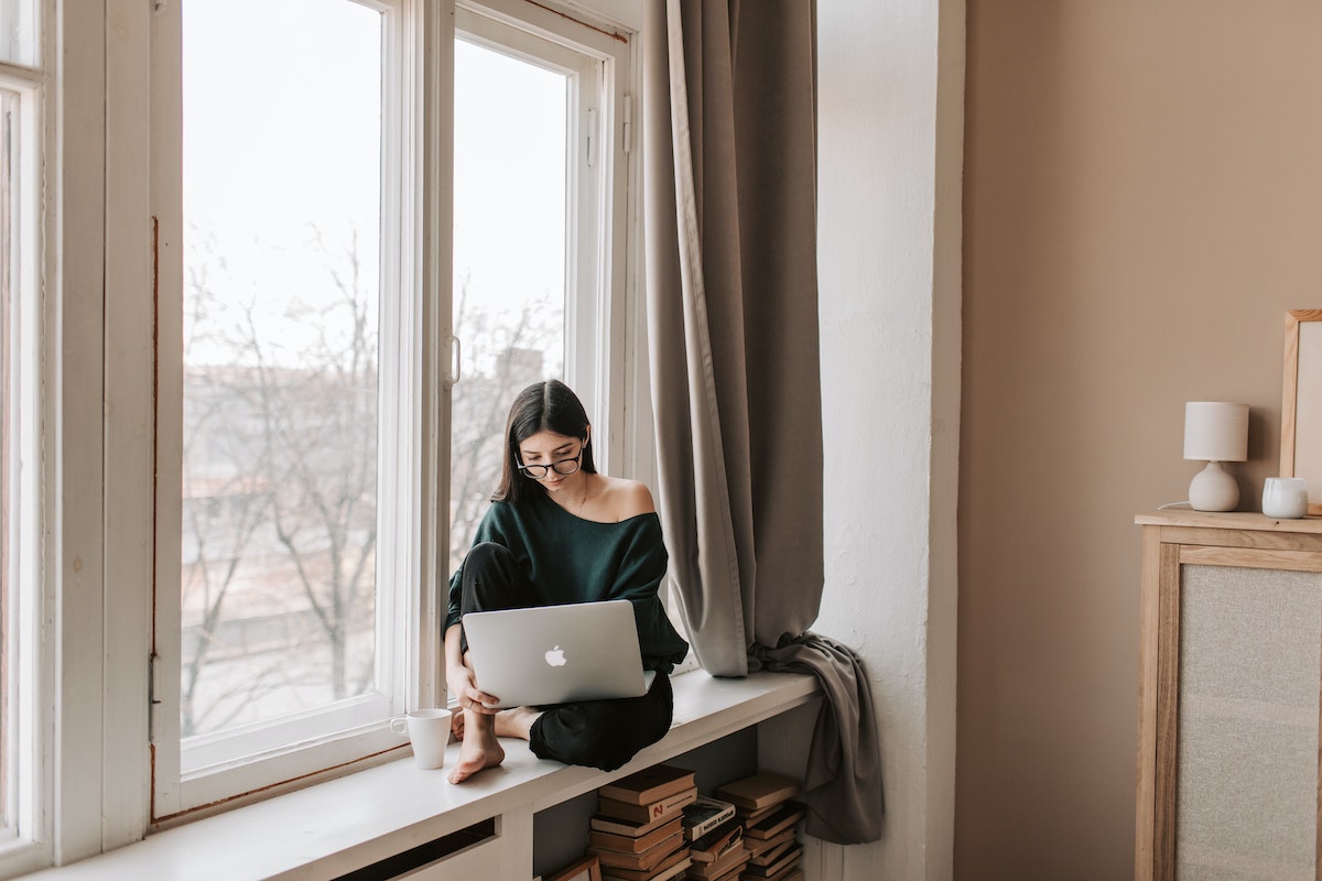 Young-female-freelancer-sitting-with-laptop-on-windowsill-at-home
